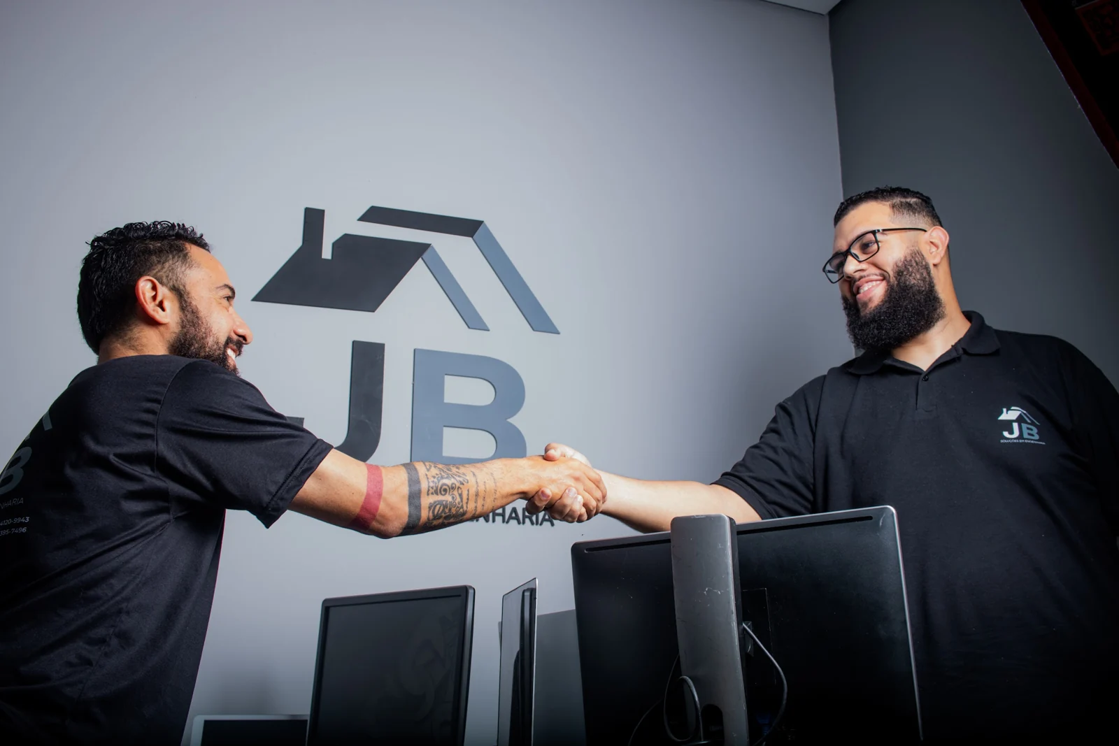 Duas pessoas se cumprimentam em um escritório, diante da marca JB Soluções em Engenharia